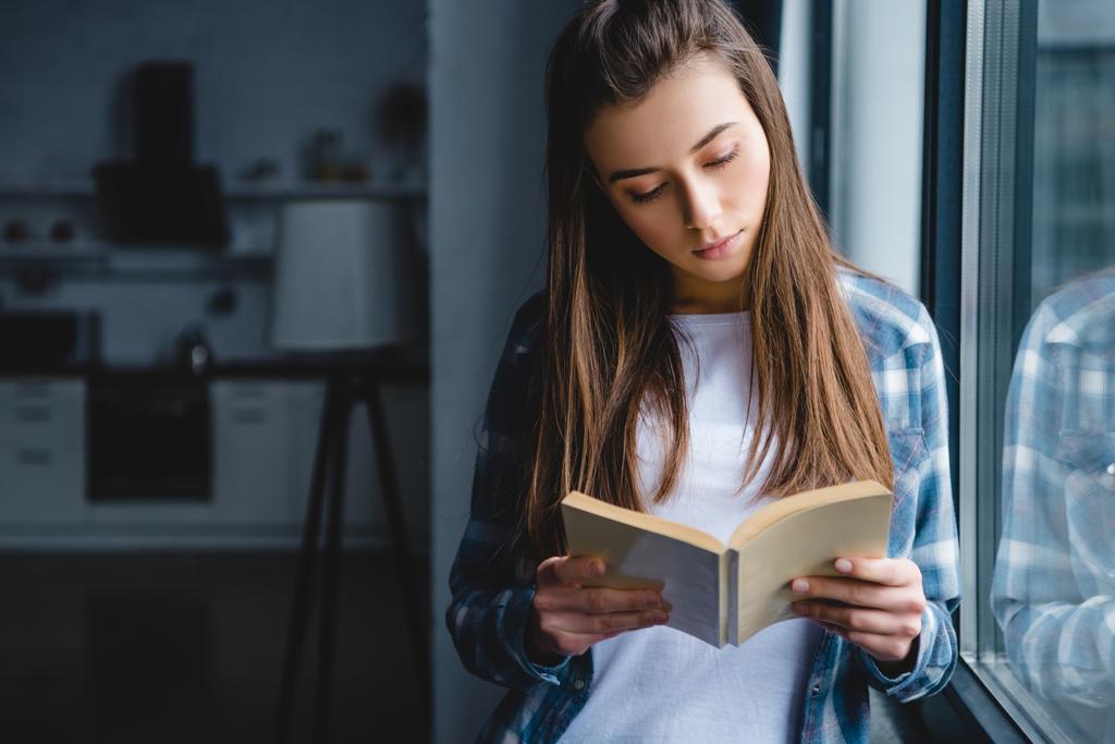 gyönyörű fiatal nő az ablak közelében, és könyvet olvas kockás ing - Fotó, kép