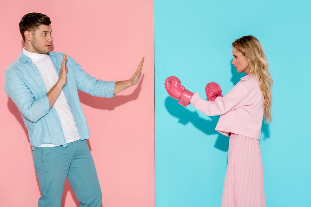 переляканий чоловік жестикулює руками біля агресивної жінки в рожевих боксерських рукавичках на рожевому і синьому фоні
 - Фото, зображення