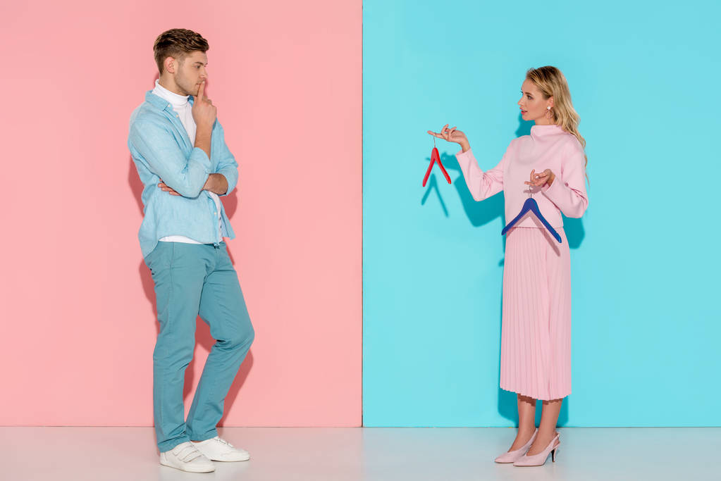 nadenkende man vrouw kijken en lege kleerhangers op roze en blauwe achtergrond te kiezen - Foto, afbeelding