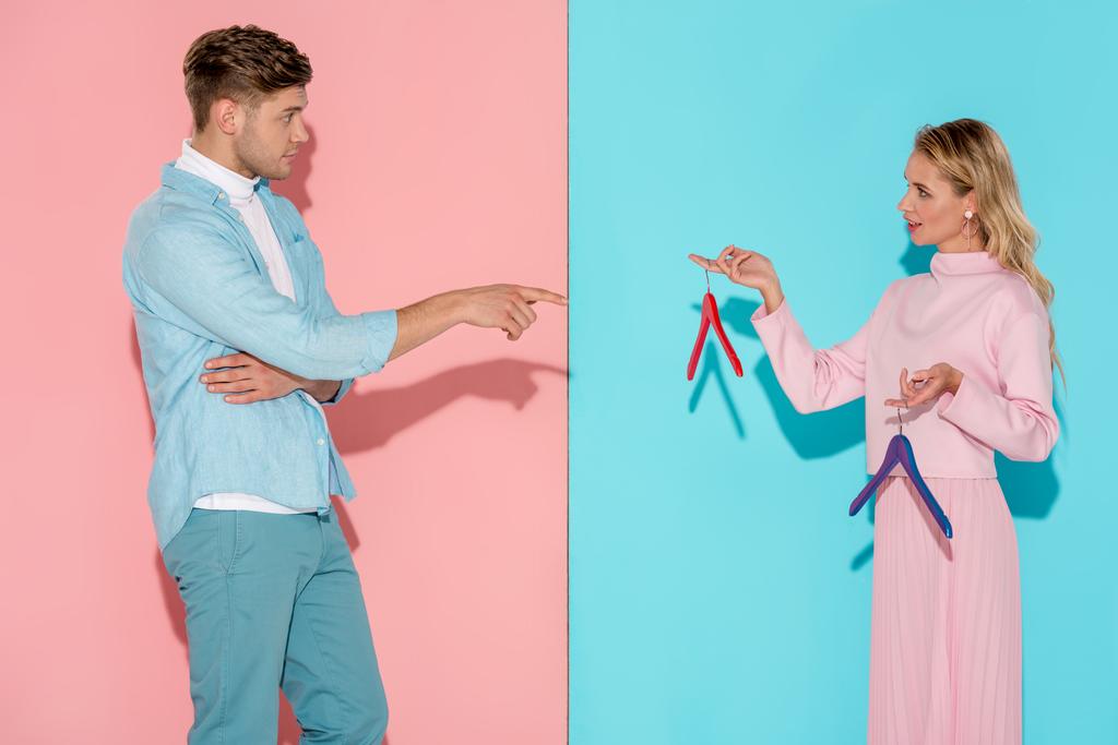handsome man pointing with finger at woman and choosing empty clothes hangers on pink and blue background - Photo, Image