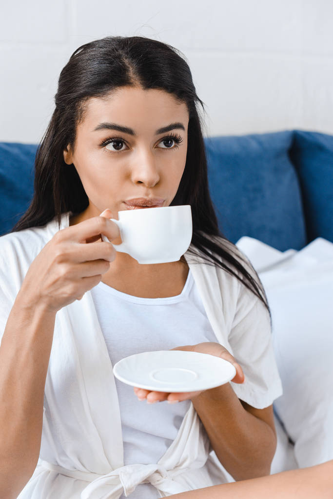 schönes Mischlingsmädchen in weißem Gewand, das morgens im Bett Kaffee trinkt - Foto, Bild
