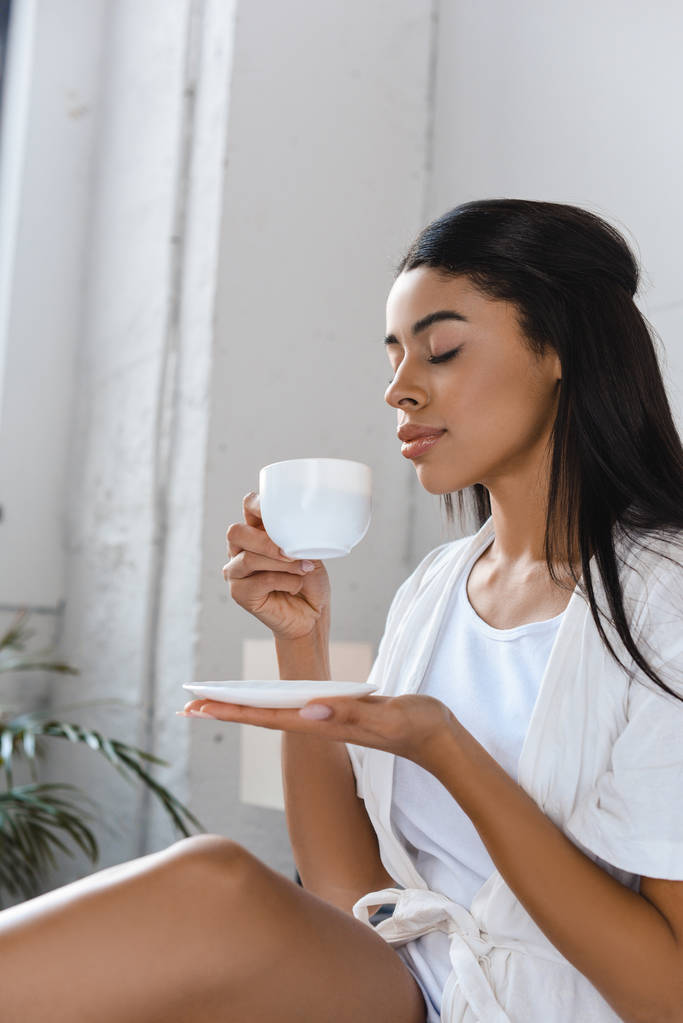 Seitenansicht der schönen gemischten Rasse Mädchen in weißen Gewand schnüffelt aromatischen Kaffee im Bett am Morgen - Foto, Bild