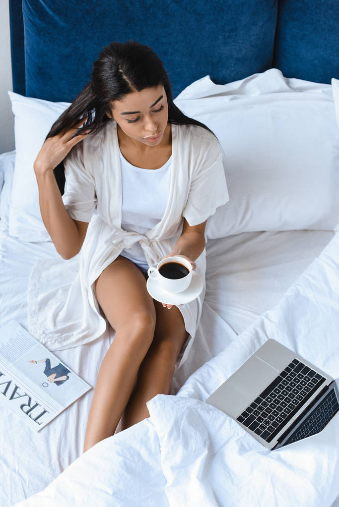 vista de ángulo alto de hermosa chica de raza mixta en túnica blanca sosteniendo taza de café en la cama por la mañana y mirando el ordenador portátil
 - Foto, imagen