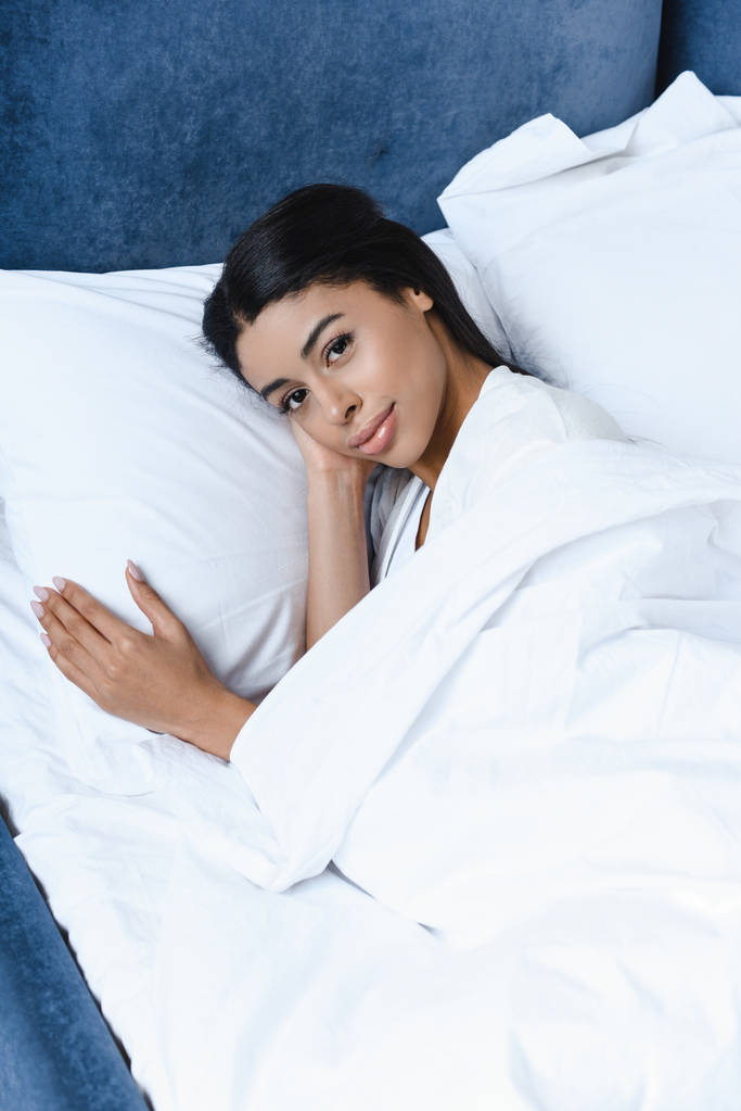 vista de ángulo alto de hermosa chica de raza mixta en bata blanca acostado en la cama por la mañana y mirando a la cámara
 - Foto, Imagen