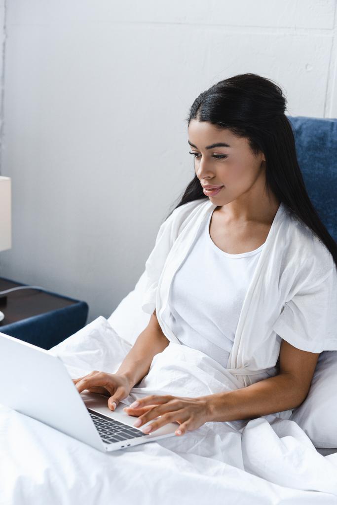 aantrekkelijke gemengd ras meisje in een witte robe met laptop in de ochtend in bed - Foto, afbeelding