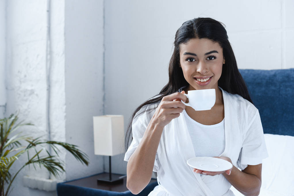 sonriente hermosa chica de raza mixta en túnica blanca beber café y mirando a la cámara en la mañana en el dormitorio
 - Foto, Imagen