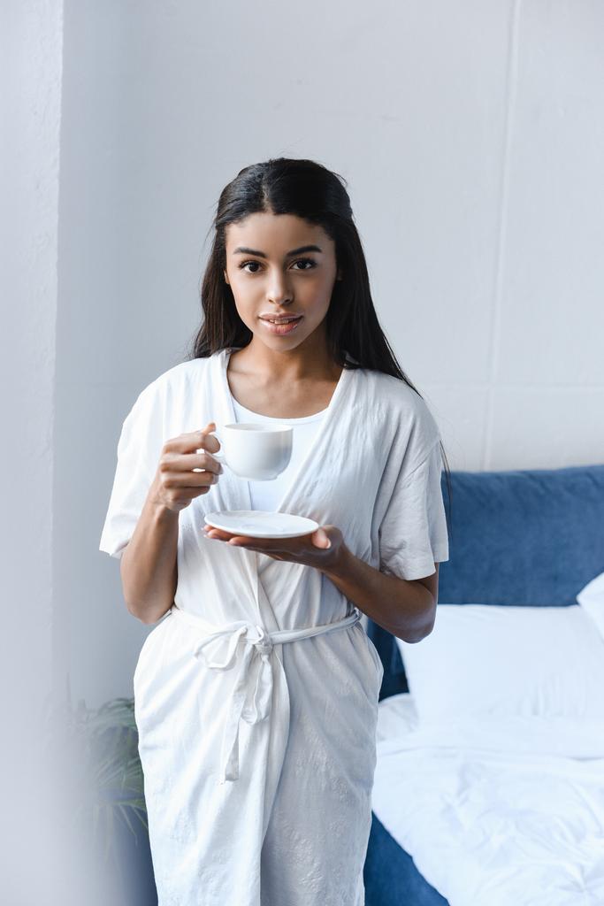 gyönyörű vegyes verseny lány fehér köntöst gazdaság csésze kávé reggel, a hálószobában, és látszó-on fényképezőgép - Fotó, kép