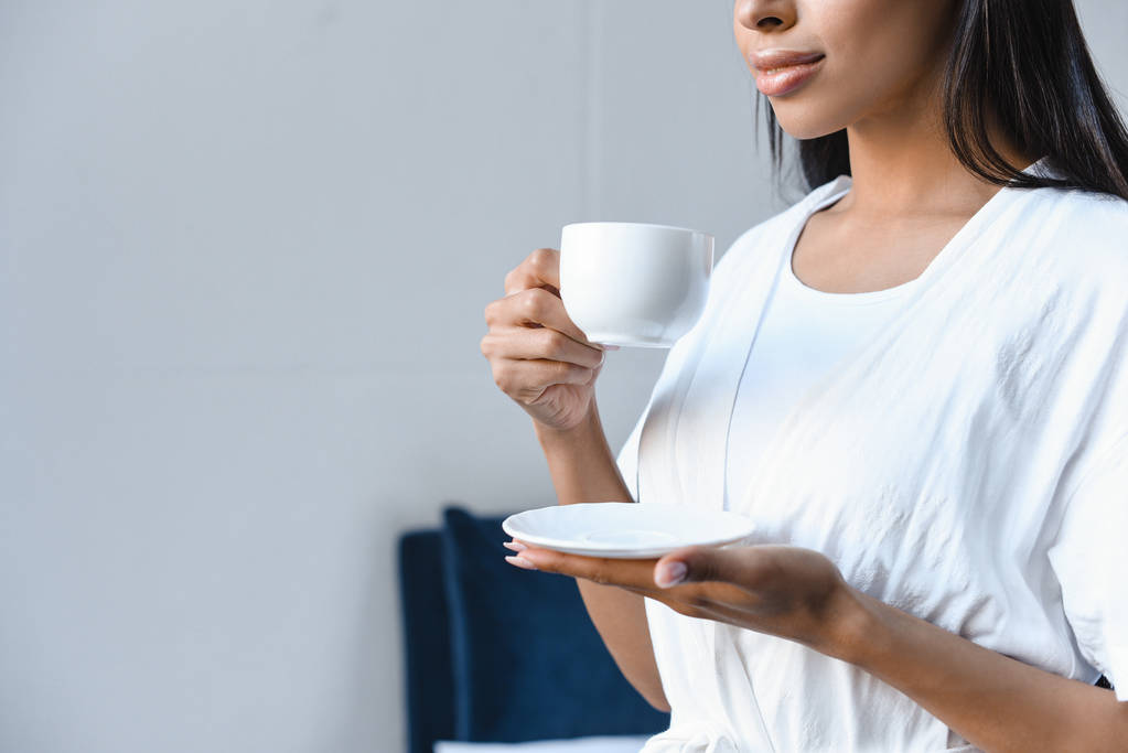 обрезанный образ смешанной расы девушка в белом халате держа чашку кофе утром в спальне
 - Фото, изображение