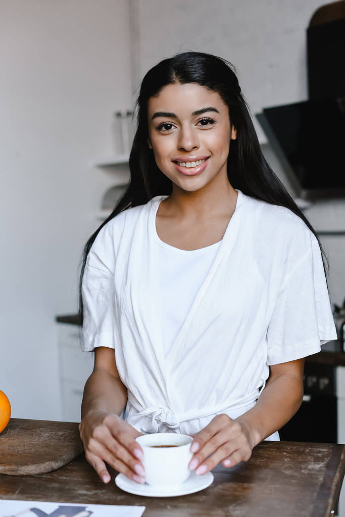 усміхнена красива змішана дівчина гонки в білому халаті тримає чашку кави і дивиться на камеру вранці на кухні
 - Фото, зображення