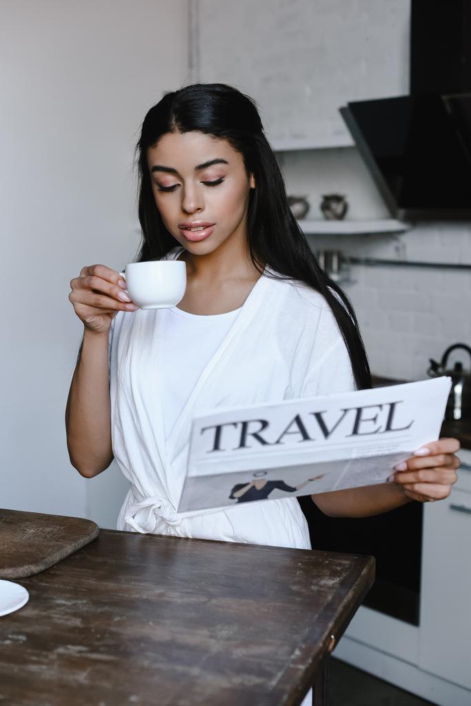 красивая смешанная раса девушка в белом халате держа чашку кофе и читать туристические газеты утром на кухне
 - Фото, изображение