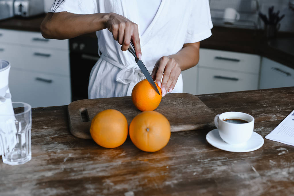 περικομμένη εικόνα της μικτής φυλή κορίτσι σε λευκό χιτώνα κοπής πορτοκάλια πρωί στην κουζίνα - Φωτογραφία, εικόνα