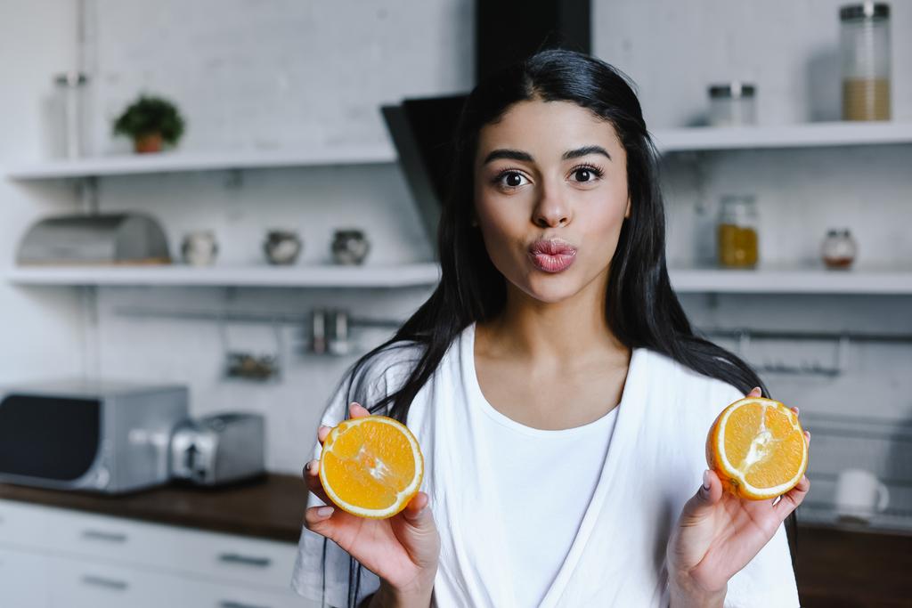 beautiful mixed race girl in white robe holding cut orange, grimacing and looking at camera in morning in kitchen - Photo, Image