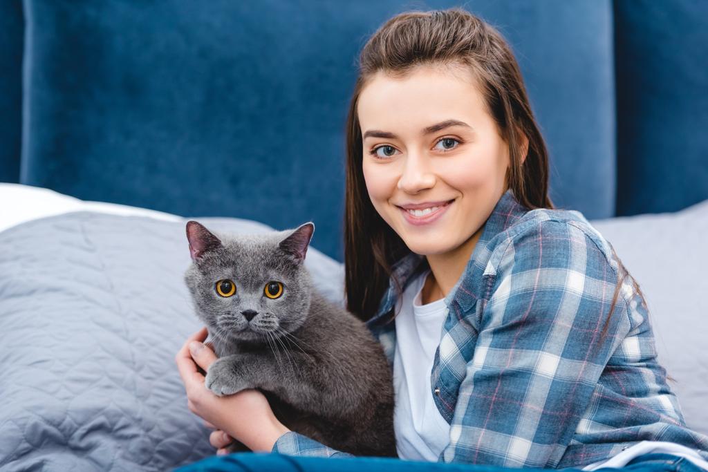 hermosa feliz joven mujer abrazando gato y sonriendo a la cámara en el dormitorio
  - Foto, imagen
