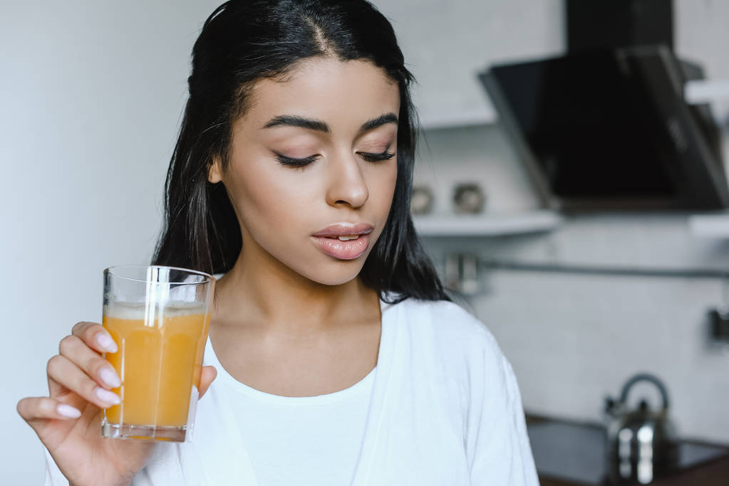 όμορφη μικτή φυλή κορίτσι σε λευκό χιτώνα, κρατά ποτήρι φρέσκο χυμό πορτοκαλιού το πρωί στην κουζίνα - Φωτογραφία, εικόνα