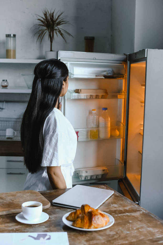 πίσω όψη της μικτής φυλή κορίτσι σε λευκό χιτώνα που ψάχνει σε ψυγείο πρωί στην κουζίνα - Φωτογραφία, εικόνα