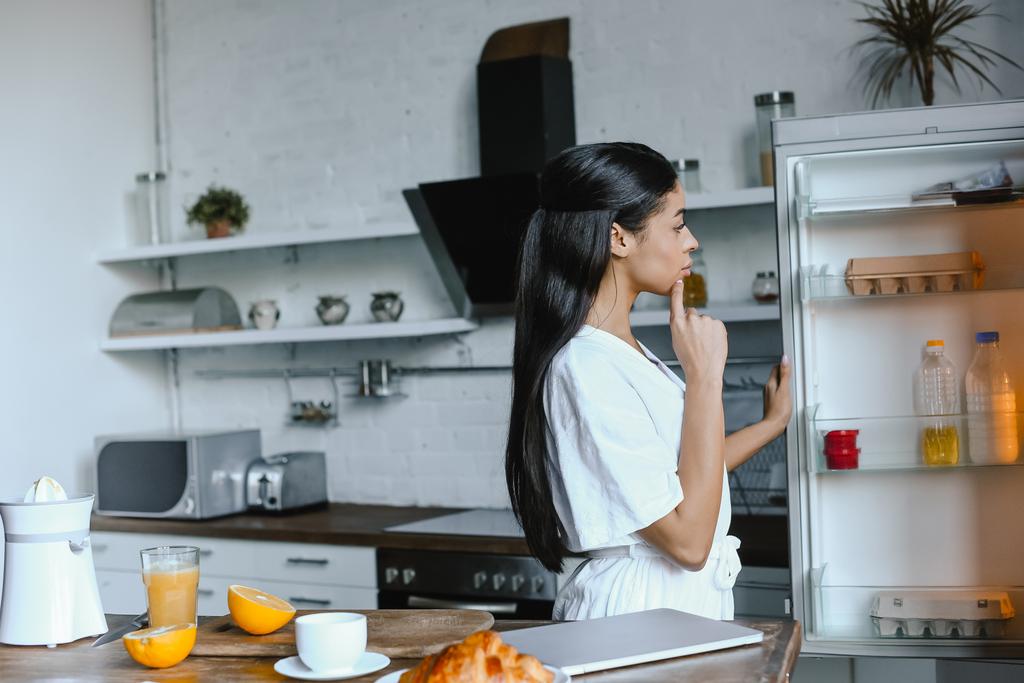 vista lateral da bela menina de raça mista pensiva em roupão branco olhando para a geladeira de manhã na cozinha
 - Foto, Imagem