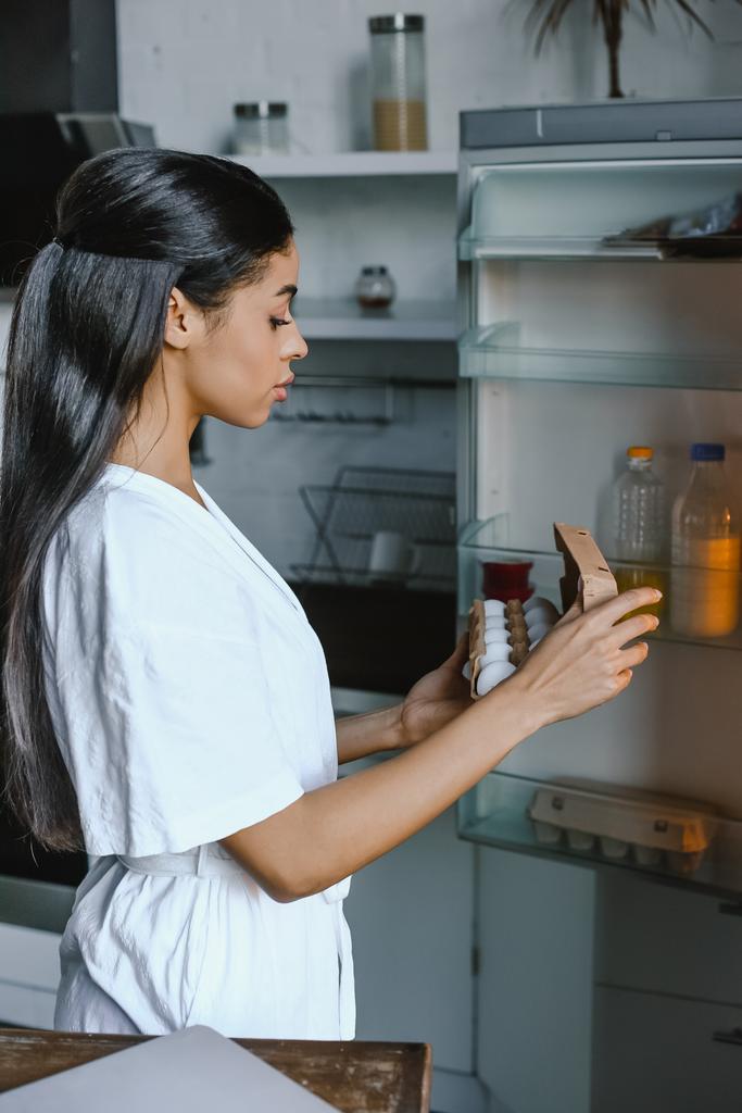 Seitenansicht der schönen Mischlingshündin in weißer Robe, die morgens in der Küche Tablett mit Eiern aus dem offenen Kühlschrank einnimmt - Foto, Bild