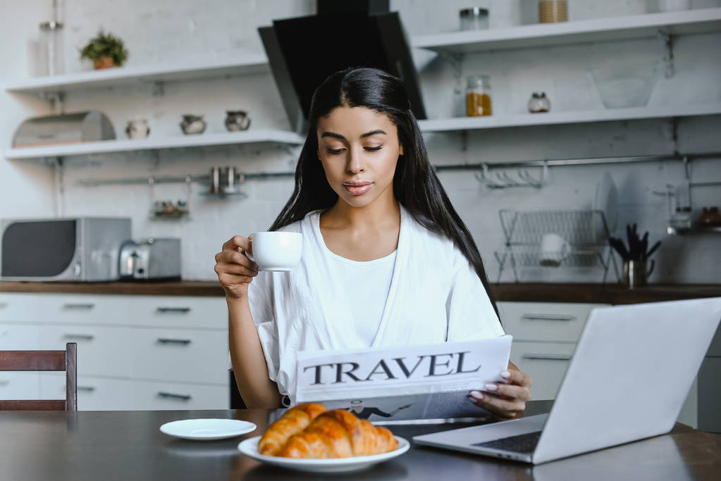 bela menina de raça mista em roupão branco segurando xícara de café e lendo jornal de viagem de manhã na cozinha
 - Foto, Imagem