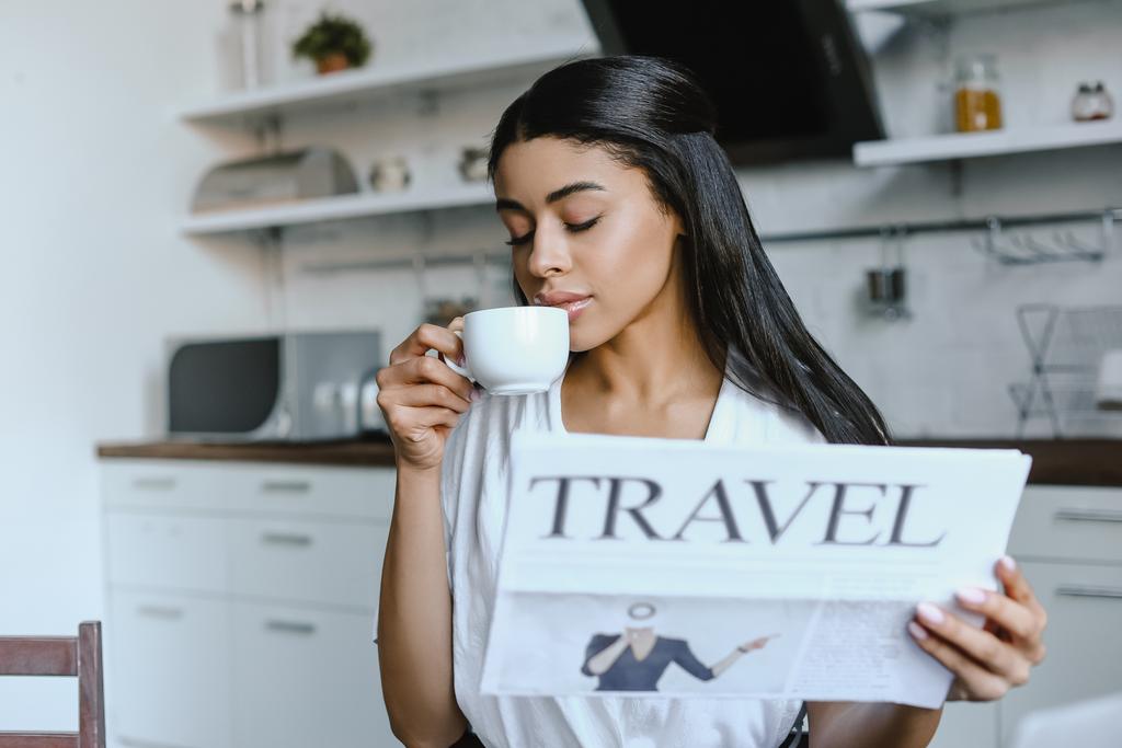 красивая смешанная раса девушка в белом халате пить кофе и проведение путешествия газеты утром на кухне
 - Фото, изображение