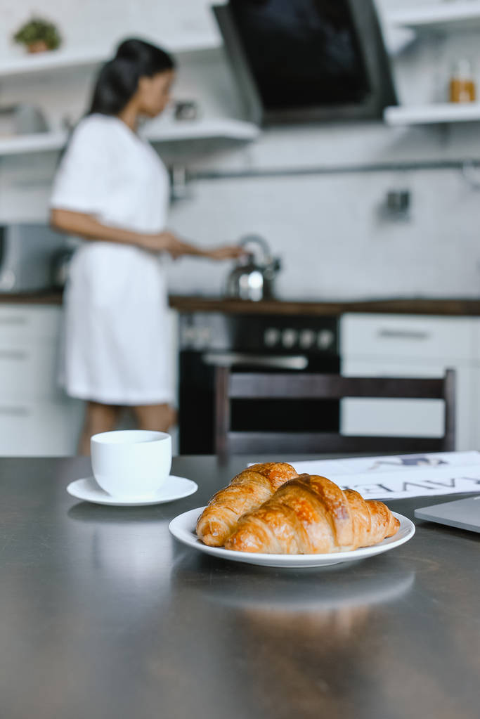 menina de raça mista em roupão branco colocando chaleira no fogão de manhã na cozinha, croissants em primeiro plano
 - Foto, Imagem