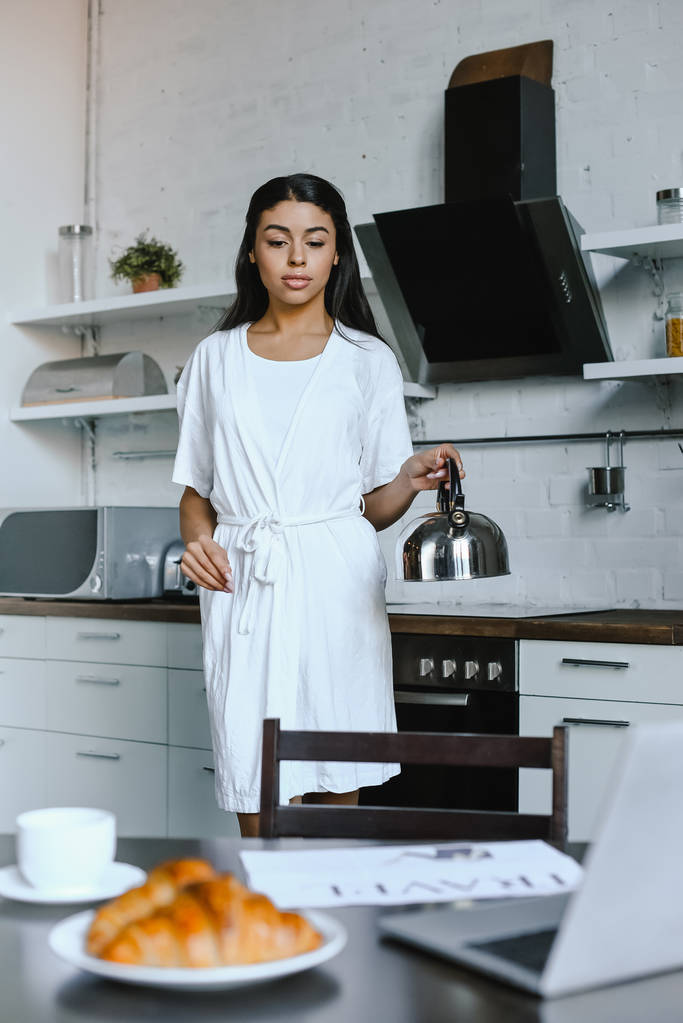 Dziewczyna piękna mieszanej rasy w białą szatę, trzymając czajnik rano w kuchni - Zdjęcie, obraz