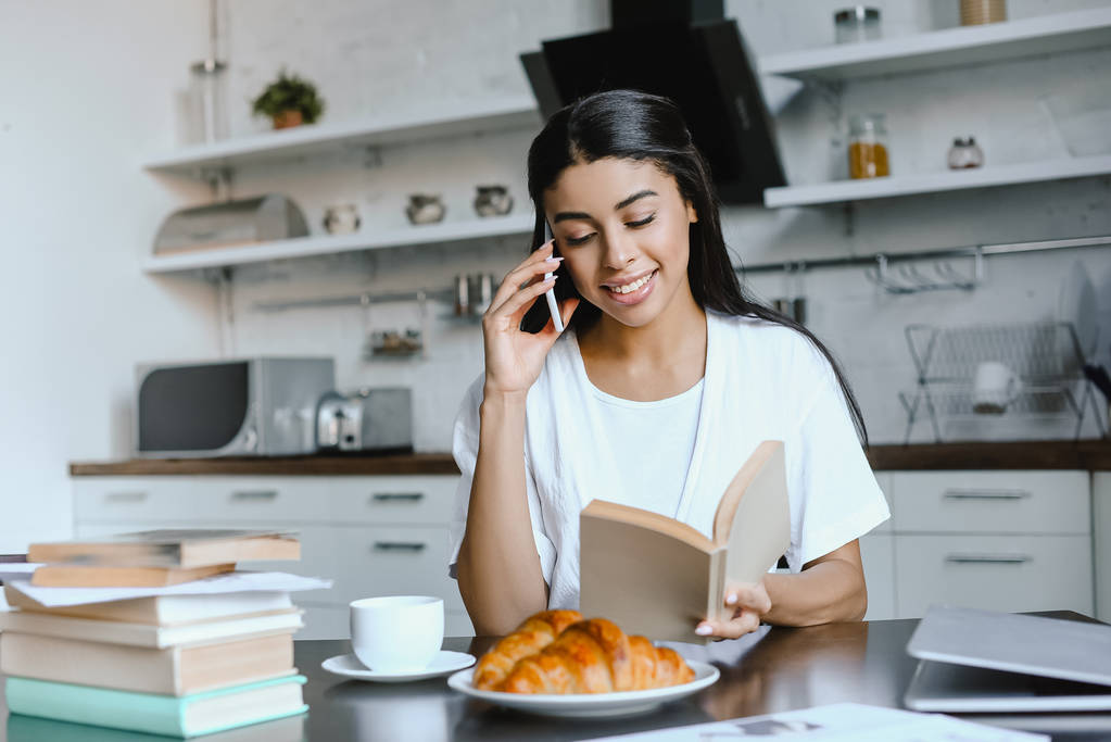lächelnde schöne Mischlingshündin in weißer Robe, die morgens in der Küche per Smartphone spricht und ein Buch in der Hand hält - Foto, Bild