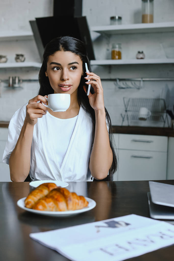 aantrekkelijke gemengd ras meisje in een witte robe praten door smartphone in de ochtend in de keuken en het drinken van koffie - Foto, afbeelding