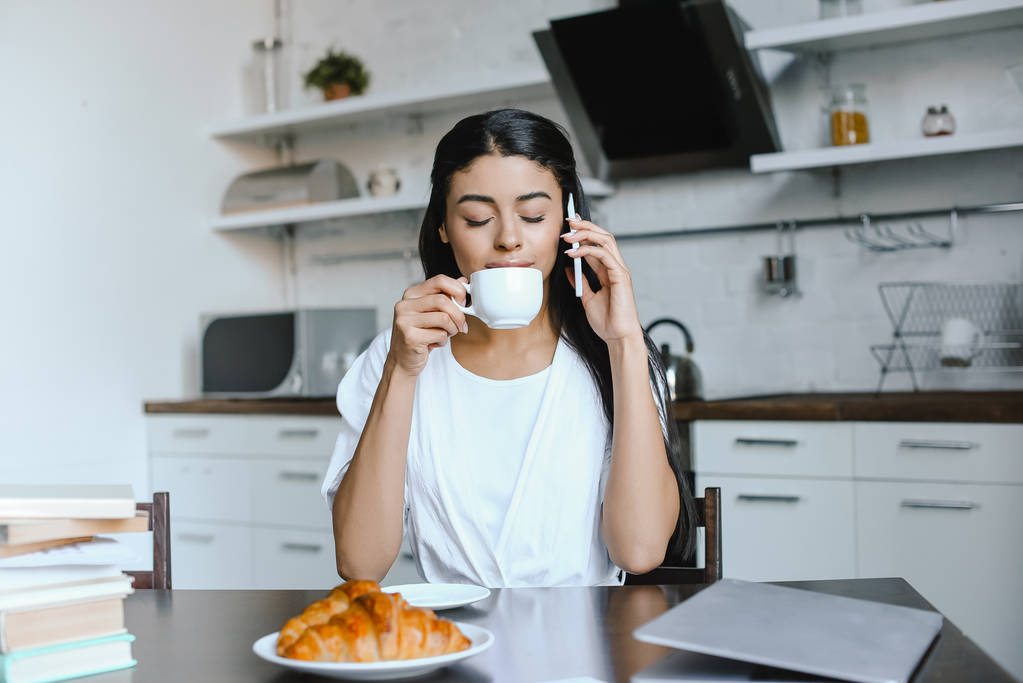hermosa chica de raza mixta en bata blanca hablando por teléfono inteligente en la mañana en la cocina y beber café con los ojos cerrados
 - Foto, Imagen