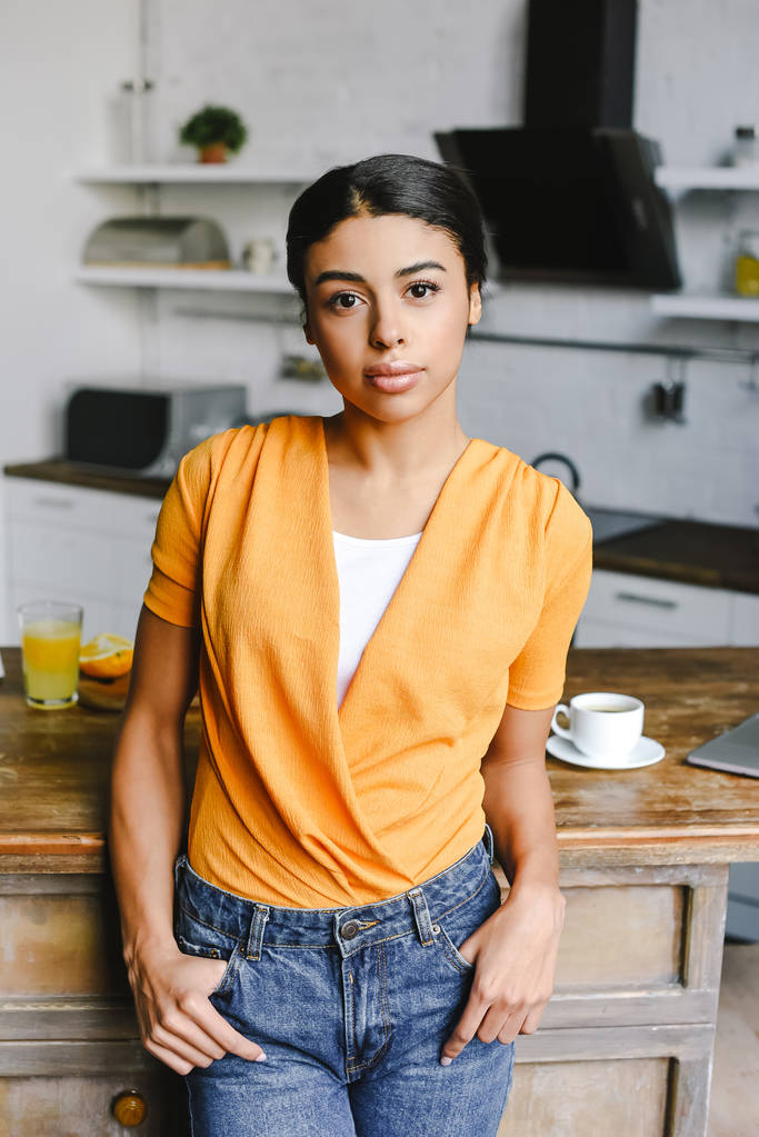 hermosa chica de raza mixta en camisa naranja de pie en la cocina y mirando a la cámara
 - Foto, imagen