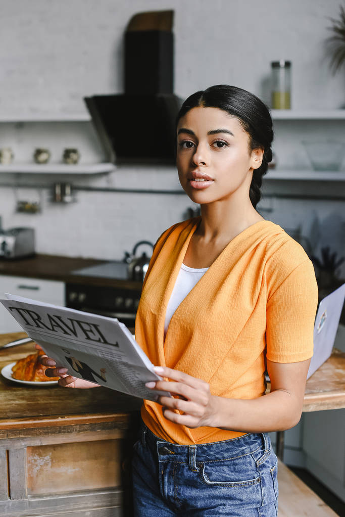 bela menina de raça mista em camisa laranja segurando jornal de viagem na cozinha e olhando para a câmera
 - Foto, Imagem
