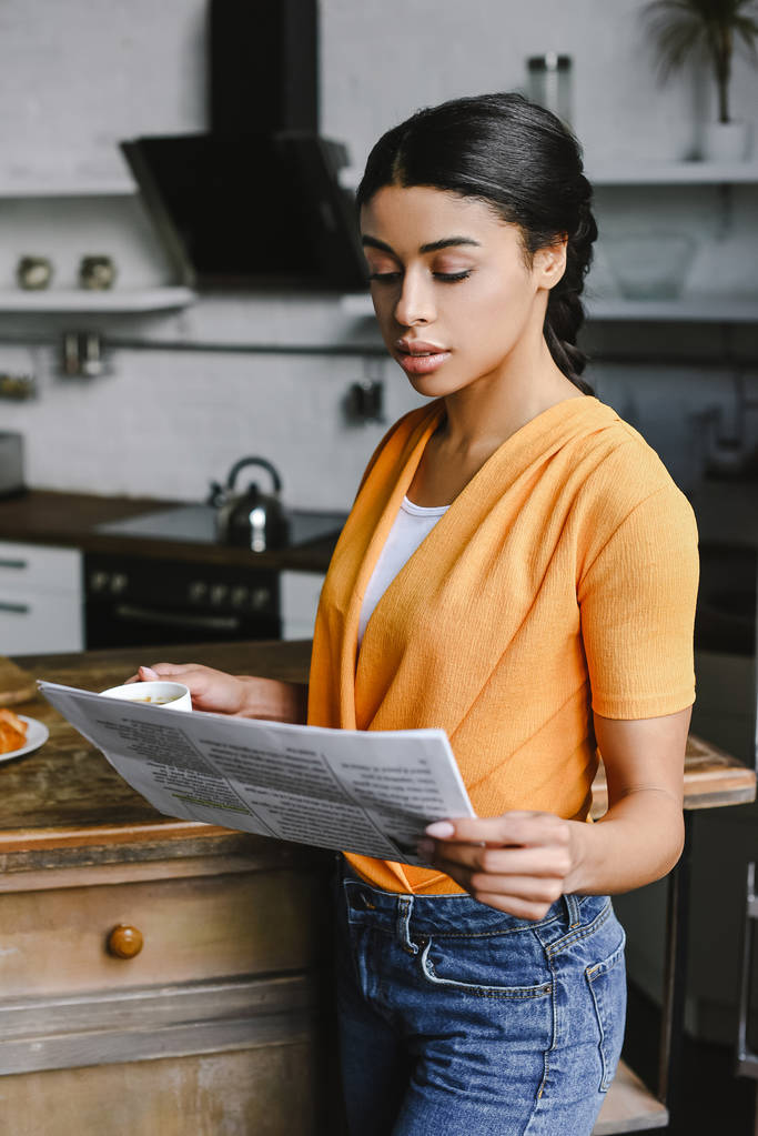 beautiful mixed race girl in orange shirt holding cup of coffee and reading newspaper in kitchen - Photo, Image