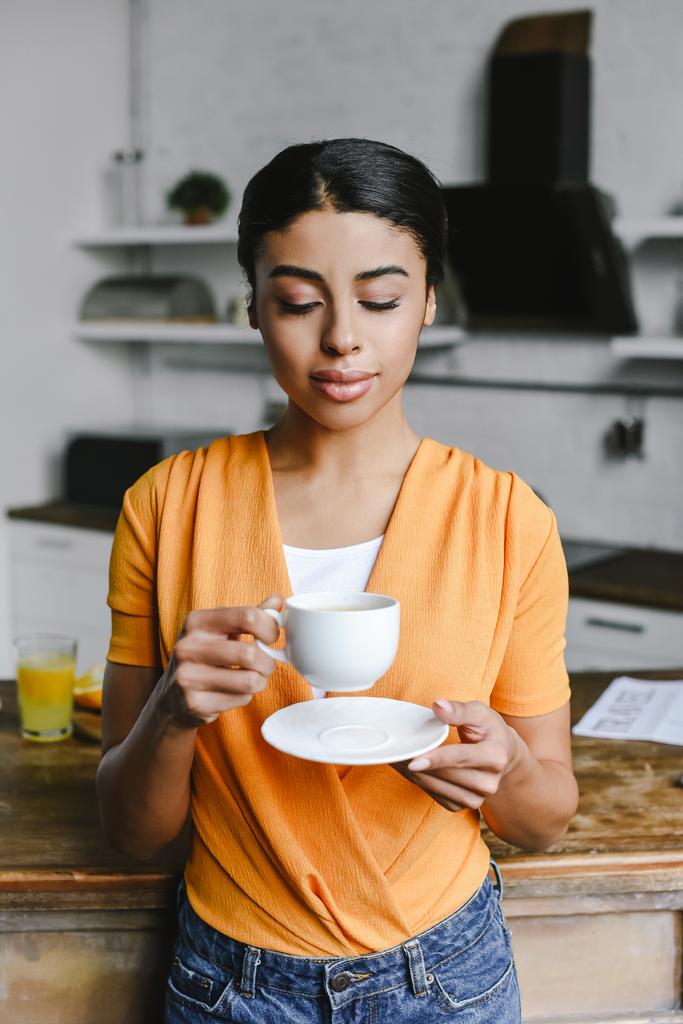 attraktives Mischlingsmädchen in orangefarbenem Hemd mit Tasse Kaffee und Teller morgens in der Küche - Foto, Bild