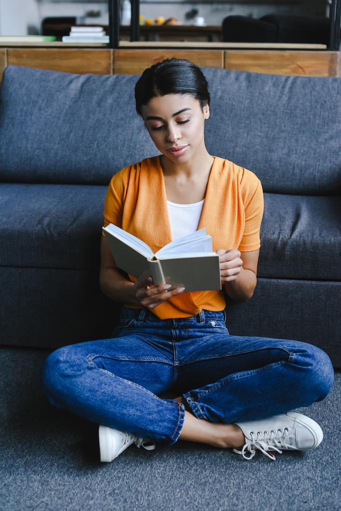 beautiful mixed race girl in orange shirt reading book on floor in living room - Photo, Image