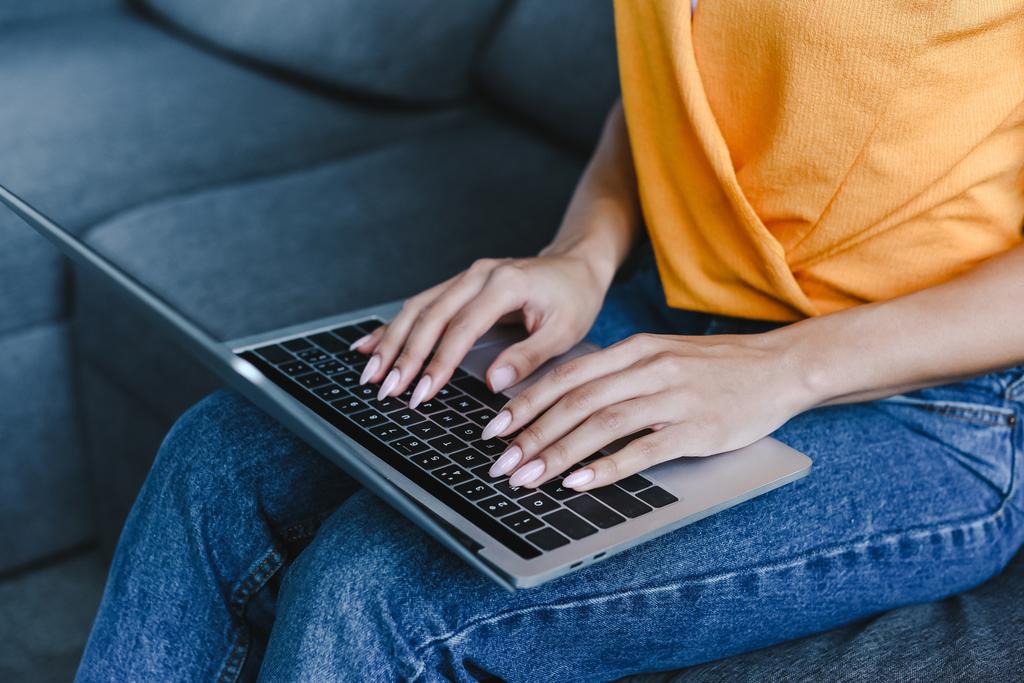 обрізане зображення змішаної гонки дівчини в помаранчевій сорочці з ноутбуком на дивані у вітальні
 - Фото, зображення