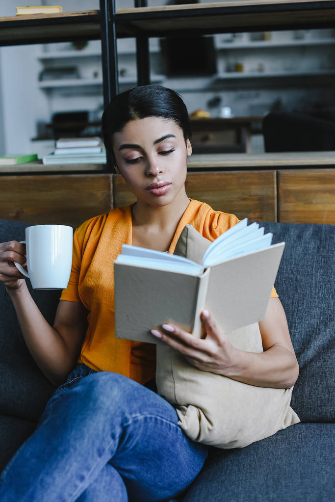 красивая смешанная раса девушка в оранжевой рубашке держа чашку чая и чтение книги на диване дома
 - Фото, изображение