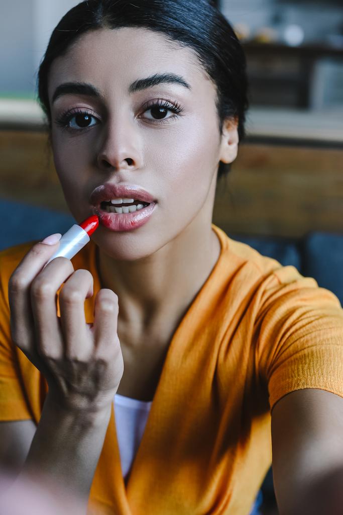 camera point of view of beautiful mixed race girl in orange shirt applying red lipstick at home and looking at camera - Photo, Image