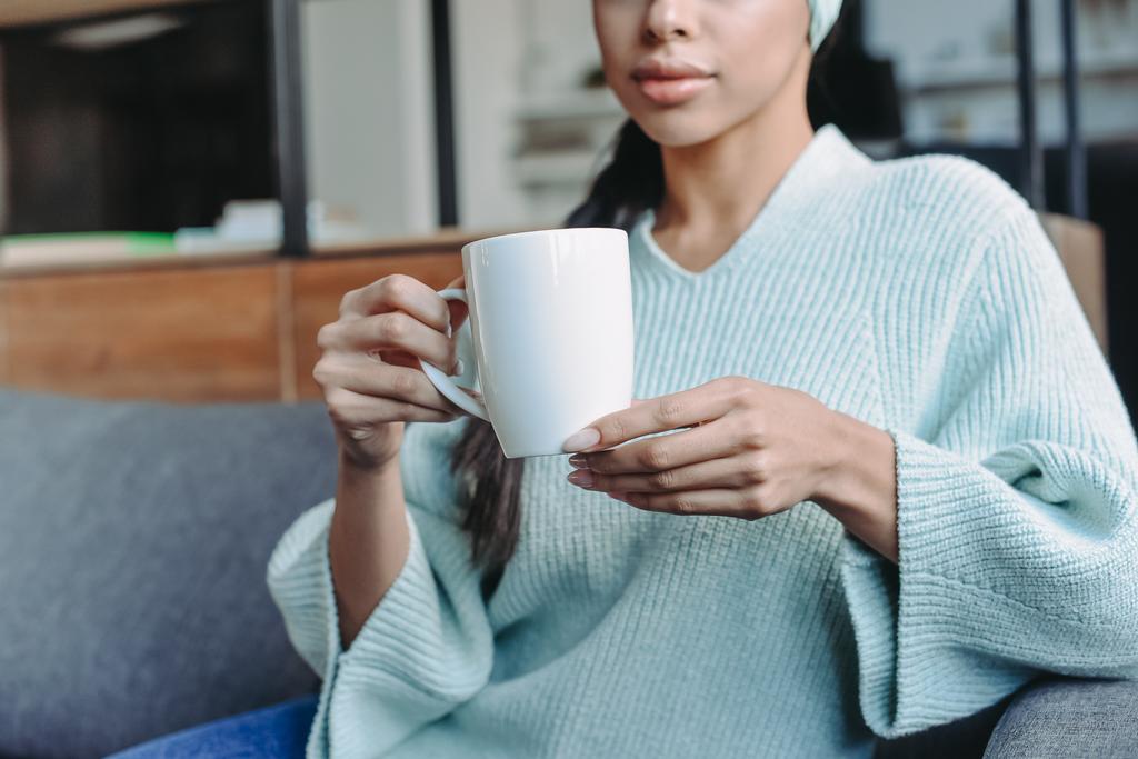 image recadrée de fille de race mixte en pull turquoise et bandeau assis sur le canapé et tenant une tasse de thé dans le salon
 - Photo, image