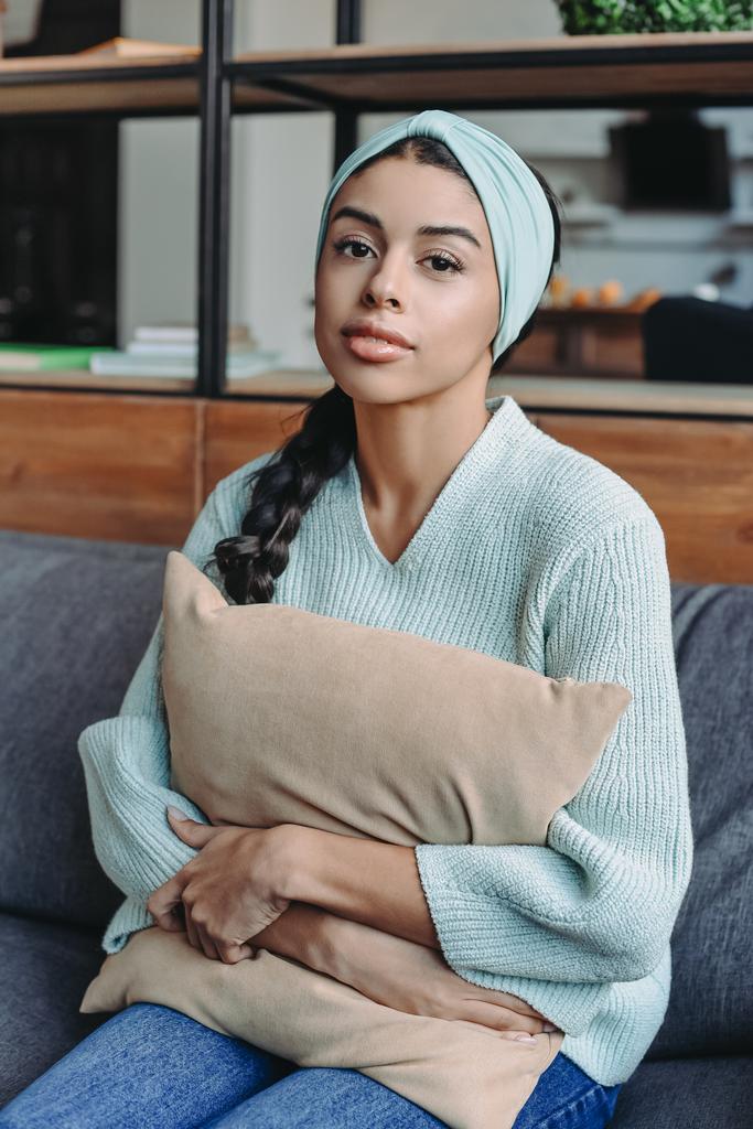 menina de raça mista atraente em camisola turquesa e almofada abraçando headband no sofá na sala de estar
 - Foto, Imagem