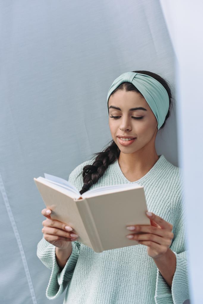 hermosa chica de raza mixta en suéter turquesa y diadema libro de lectura en casa
 - Foto, imagen