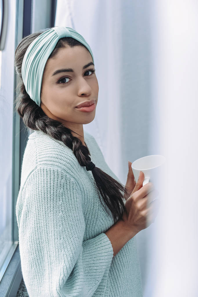 gyönyörű kevert lány türkizkék pulóver és fejpánt otthon tartja a csésze teát, és látszó-on fényképezőgép - Fotó, kép