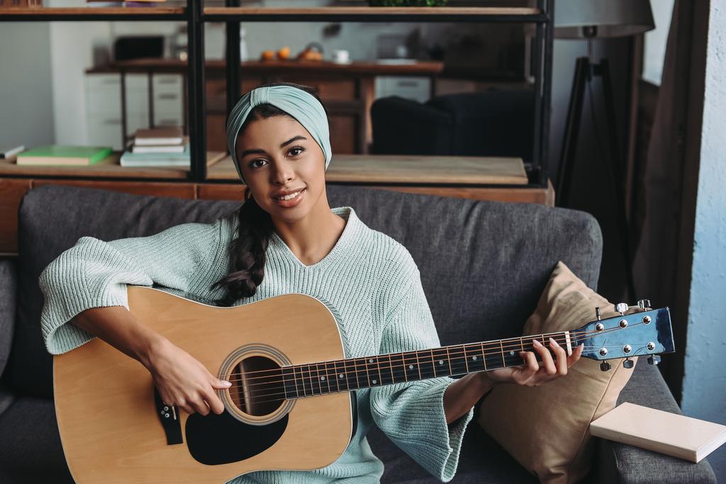 aantrekkelijke gemengd ras meisje in turkooizen trui en hoofdband akoestische gitaar spelen op sofa thuis en kijken naar camera - Foto, afbeelding