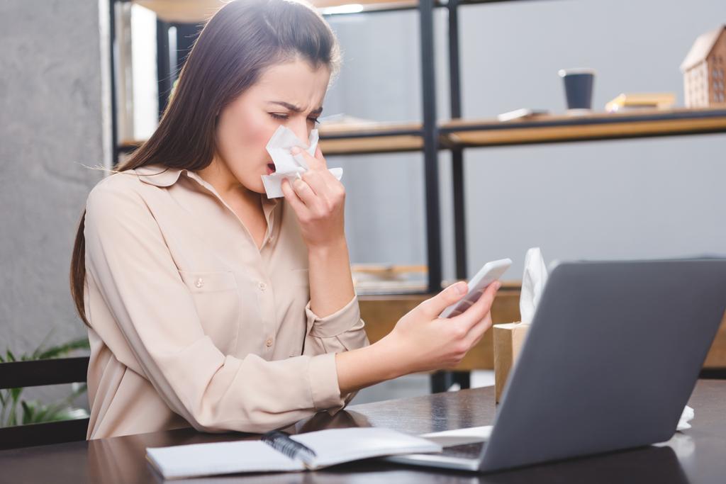 jonge zakenvrouw holding smartphone en lijden aan allergie op werkplek   - Foto, afbeelding
