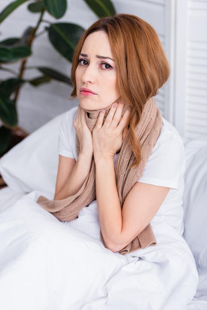 Kranke Frau berührt schmerzenden Hals mit Schal und sitzt zu Hause auf Bett und blickt in Kamera - Foto, Bild