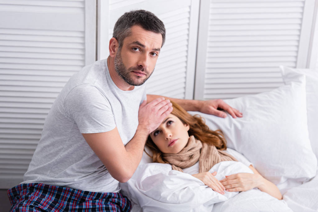 Красивый муж трогает лоб больной жены с лихорадкой в постели, смотрит в камеру
 - Фото, изображение