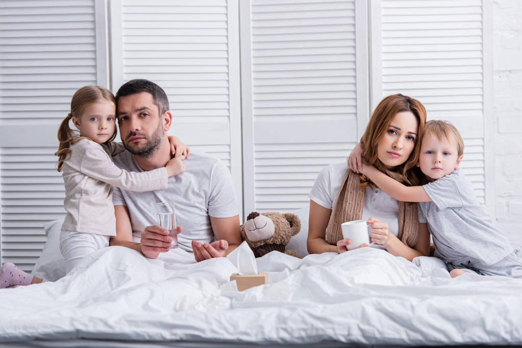 adorable dzieci przytulanie chore matki i ojca w sypialni, patrząc na kamery - Zdjęcie, obraz