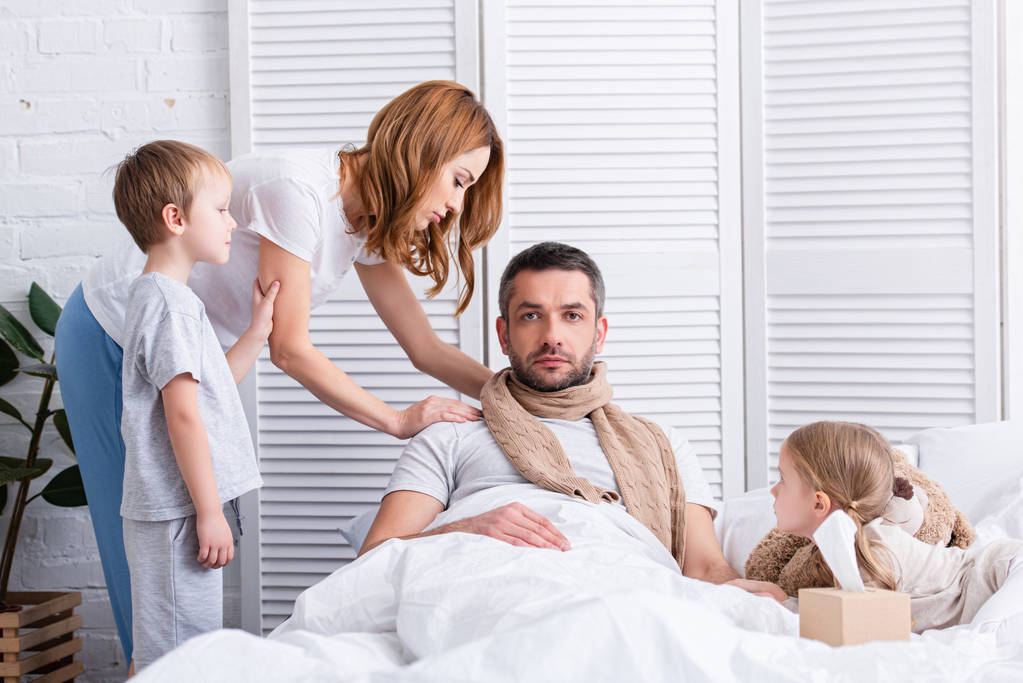 vaimo ja lapset huolehtivat sairaasta isästä makuuhuoneessa, hän makaa sängyssä ja katselee kameraa
 - Valokuva, kuva
