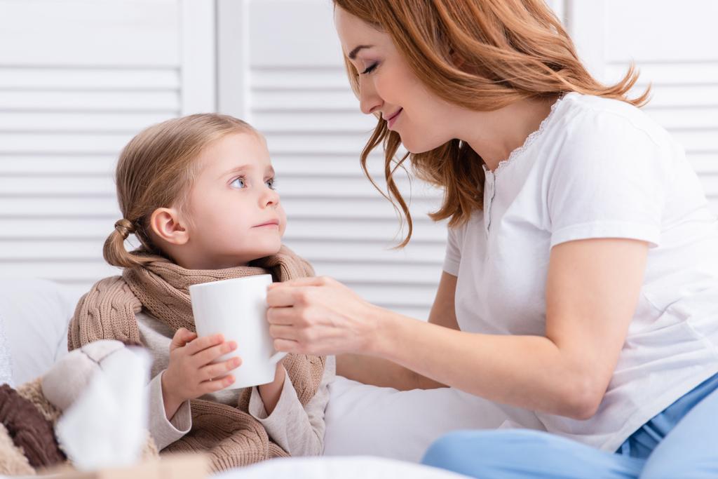 mother taking care of sick daughter and giving her cup of tea in bedroom - Photo, Image