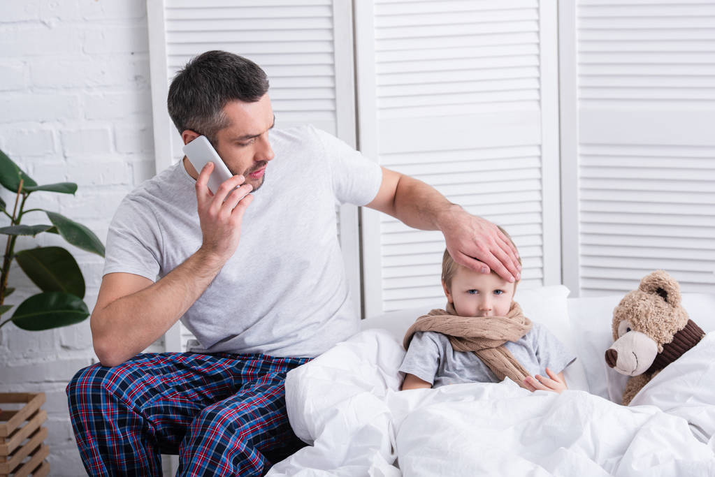 isä puhuu älypuhelimella ja koskettaa sairas poika otsa makuuhuoneessa
 - Valokuva, kuva