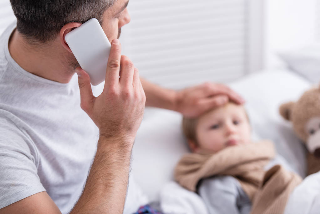 enfoque selectivo del padre hablando por teléfono inteligente y tocando la frente del hijo enfermo en el dormitorio
 - Foto, Imagen