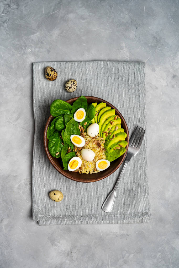 Top view of salad with bulgur, spinach, avocado and quail eggs in wooden bowl on grey linen towel - Photo, Image